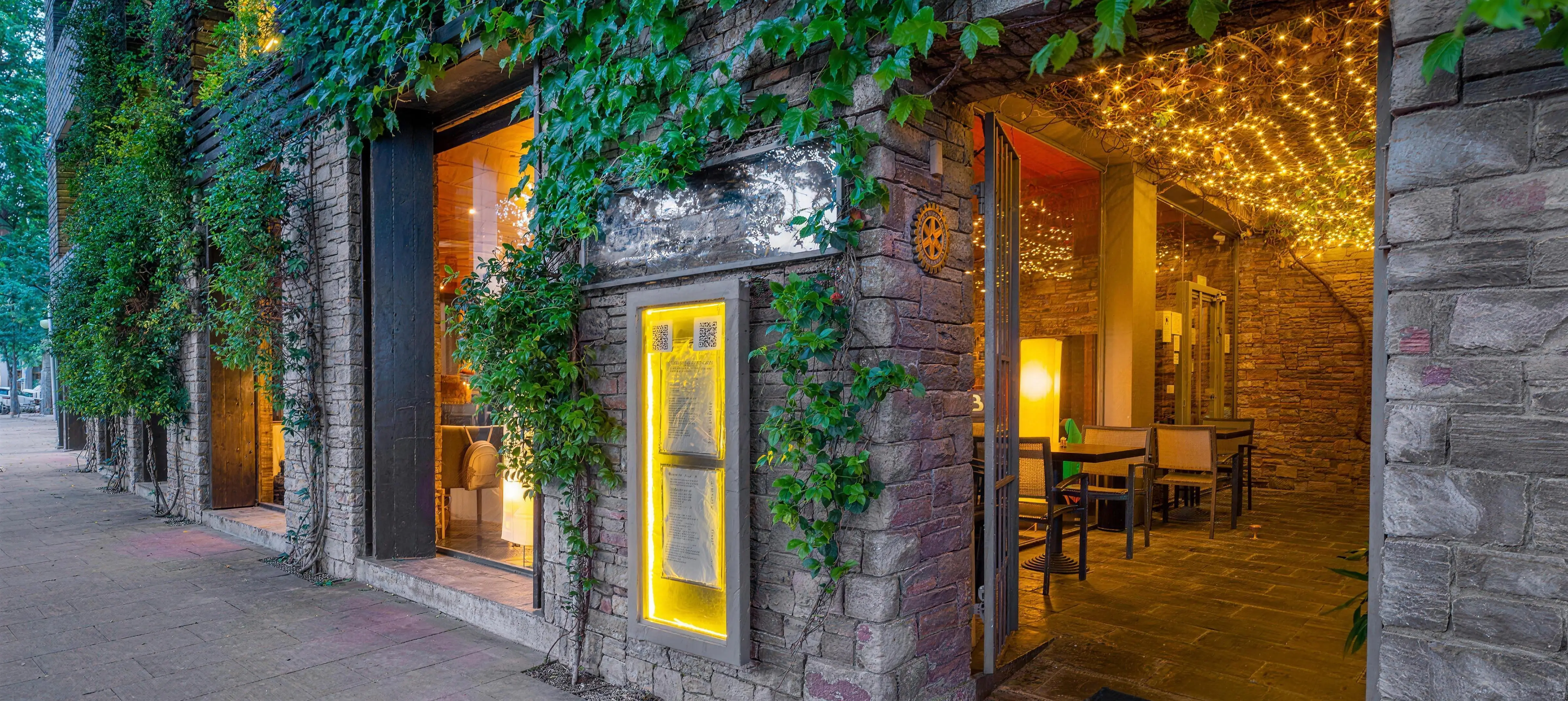 La façana del restaurant al capvespre, amb la pedra i l’heura il·luminades i les taules de dins ja parades.