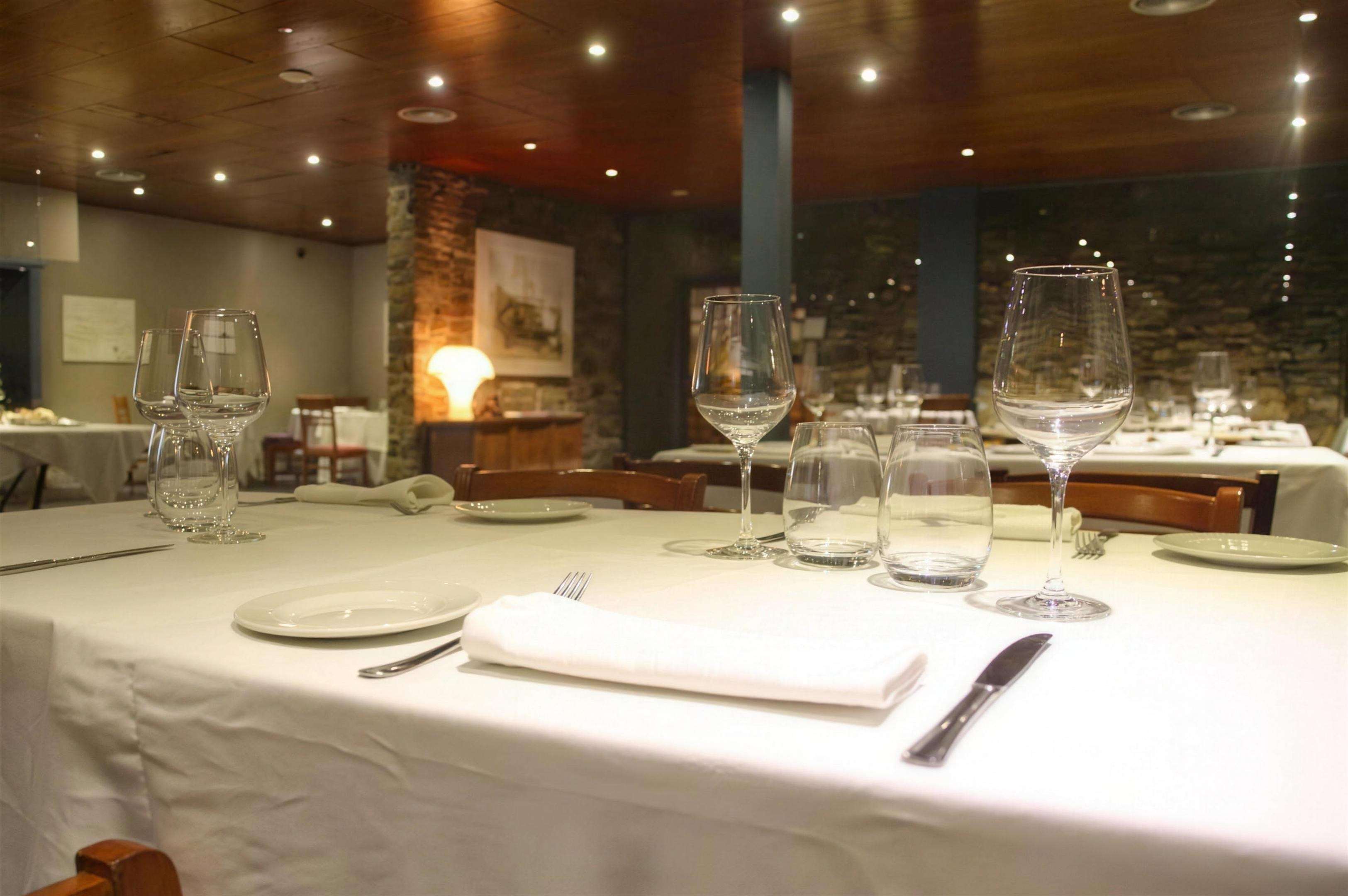 Una mesa puesta en primer término, con la sala iluminada al fondo y el techo de madera.